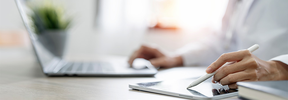 Person using a stylus on a tablet next to an open laptop on a desk, with a blurred plant in the background.